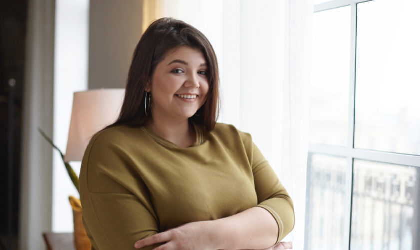 people-body-positivity-lifestyle-concept-indoor-image-adorable-overweight-chubby-young-lady-relaxing-home-standing-window-crossing-arms-her-chest-smiling-broadly